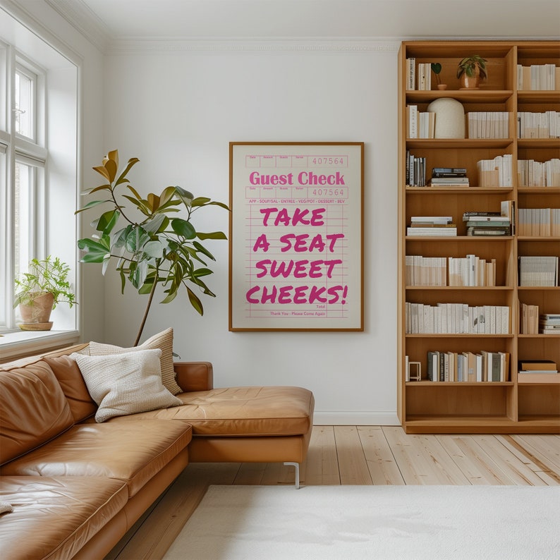 K&ouml;nnte beinhalten: Ein rosa-wei&szlig;es Poster mit dem Text "Guest Check" und "Take a seat sweet cheeks!" auf einem karierten Hintergrund. Das Poster h&auml;ngt an einer wei&szlig;en Wand in einem Wohnzimmer mit einem braunen Ledersofa und einem B&uuml;cherregal aus Holz.