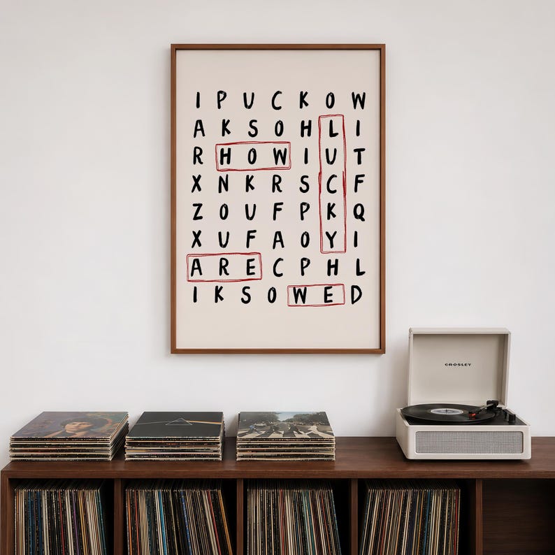 May include: A framed wall art print featuring a word search puzzle on a cream background with black letters. The words HOW, LUCKY, ARE, and WED are highlighted with red rectangular outlines. The print is enclosed in a thin brown wooden frame.