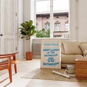May include: A framed print with the text "Guest Check" and "What Happens in the Bathroom Stays in the Bathroom" in blue on a cream background. The print is displayed in a living room setting with a window and a plant.