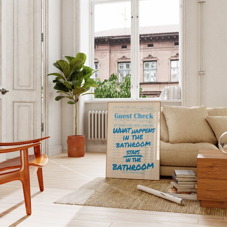May include: A framed print with the text "Guest Check" and "What Happens in the Bathroom Stays in the Bathroom" in blue on a cream background. The print is displayed in a living room setting with a window and a plant.