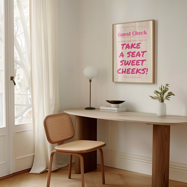 K&ouml;nnte beinhalten: Ein gerahmter Druck mit wei&szlig;em Hintergrund und rosa Text, der "Guest Check" und "Take a seat sweet cheeks!" lautet. Der Druck h&auml;ngt an einer wei&szlig;en Wand &uuml;ber einem Holztisch mit einem geflochtenen Stuhl und einer wei&szlig;en Lampe.