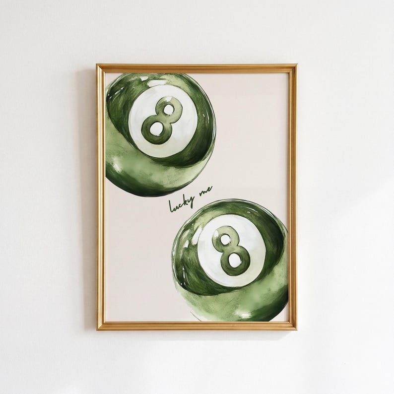 May include: A framed print featuring two green and white eight-ball illustrations. The watercolor-style artwork has a gold frame and the words "lucky me" in cursive. The background is a light pink.