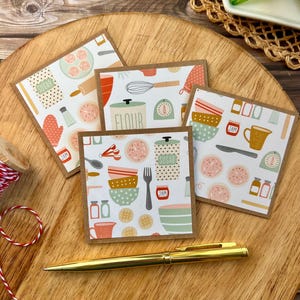 May include: Four square greeting cards with a kitchen theme, featuring illustrations of baking tools, bowls, and jars. The cards are arranged on a wooden surface with a gold pen and red and white twine.
