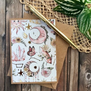 May include: A white card with a pink and gold illustration of crystals, moons, stars, butterflies, and flowers. The card is on a brown envelope.