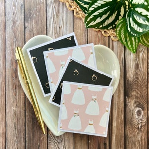 May include: A stack of four square cards featuring alternating patterns of white wedding dresses on a light pink background and gold engagement rings on a black background, accompanied by a gold pen on a heart-shaped white ceramic dish.