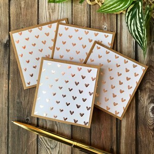 May include: Four white cards with a repeating pattern of rose gold hearts. The cards are stacked on top of each other and have a brown border.