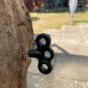 May include: A black, three-holed wind-up key with a metallic silver shaft, embedded into the side of a rough, textured wooden post. The key features a smooth, matte finish and a distinct shape with circular cutouts in each of its three lobes.