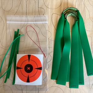 Puede incluir: Un conjunto de gomas verdes para una honda, un cordel rojo y un conjunto de gomas verdes en una bolsa de plástico transparente. También hay un blanco rojo y negro con fondo blanco.