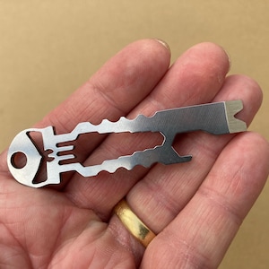 May include: A small, silver multi-tool held in a hand. The tool has a key-like shape with various cutouts and a flathead screwdriver end. The tool is made of metal and has a matte finish. The background is a neutral tan color.