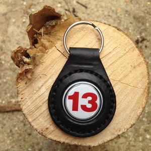 May include: Black leather keychain with a silver ring and a white circle with the number 13 in red.