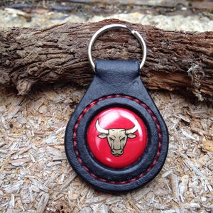 May include: Black leather keychain with a red circle featuring a gold bull head design. The keychain has a silver metal ring.