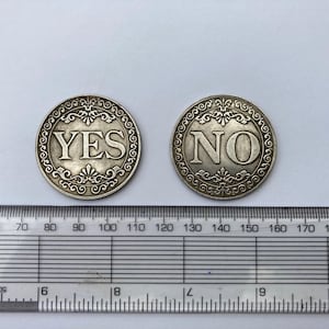 Puede incluir: Dos monedas de decisión plateadas, una con "YES" y la otra con "NO" en letras negritas. Cada moneda tiene un borde ornamental y decorativo. Una regla se coloca debajo.
