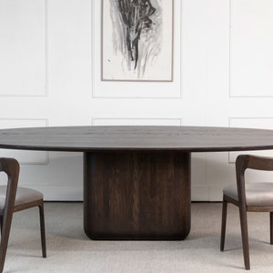 May include: A dark brown oval dining table with a solid base, flanked by two wooden chairs with gray upholstered seats. The table is approximately 108 inches long. The chairs have a curved back design. A piece of abstract art hangs on the white wall.