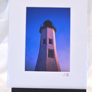 May include: A framed photograph of a white lighthouse against a blue sky. The lighthouse has a black top and two windows. The photograph is displayed on a black easel.