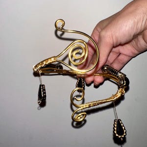 May include: Gold-toned wire jewelry with intricate swirls and black beaded accents. The piece has a unique, artistic design with dangling black beads. The jewelry is held by a hand, showcasing its delicate craftsmanship and elegant style.