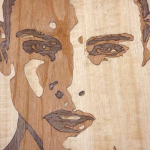 May include: A wooden portrait of a face, crafted from different wood tones. The artwork has a light-coloured background, with darker wood used to define the facial features, creating a strong contrast. This is a unique piece of art.