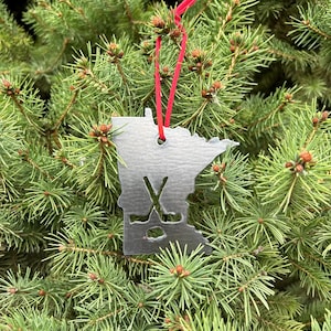 May include: A silver metal Christmas ornament shaped like the state of Minnesota with hockey sticks and a puck inside the outline. The ornament is hanging from a red ribbon on a green evergreen tree.