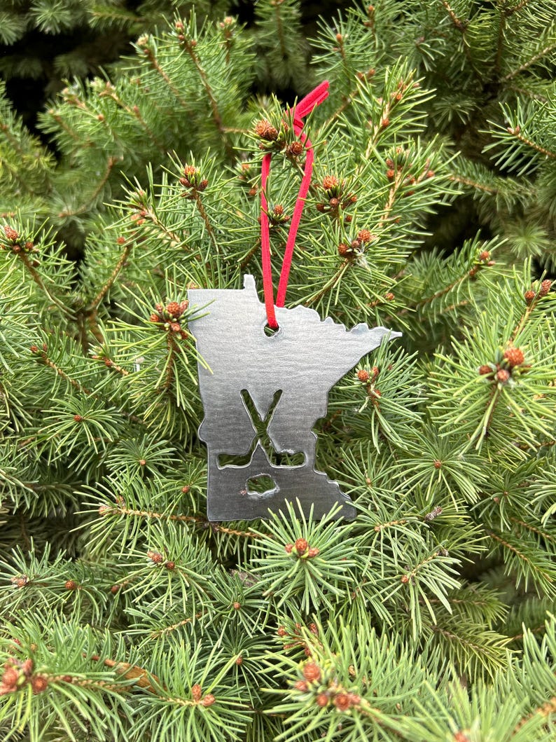 May include: A silver metal Christmas ornament shaped like the state of Minnesota with hockey sticks and a puck inside the outline. The ornament is hanging from a red ribbon on a green evergreen tree.