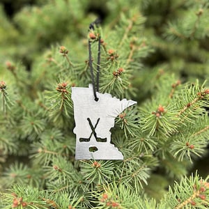 May include: A silver metal ornament shaped like the state of Minnesota with a hockey stick and puck design. The ornament is hanging on a green Christmas tree.