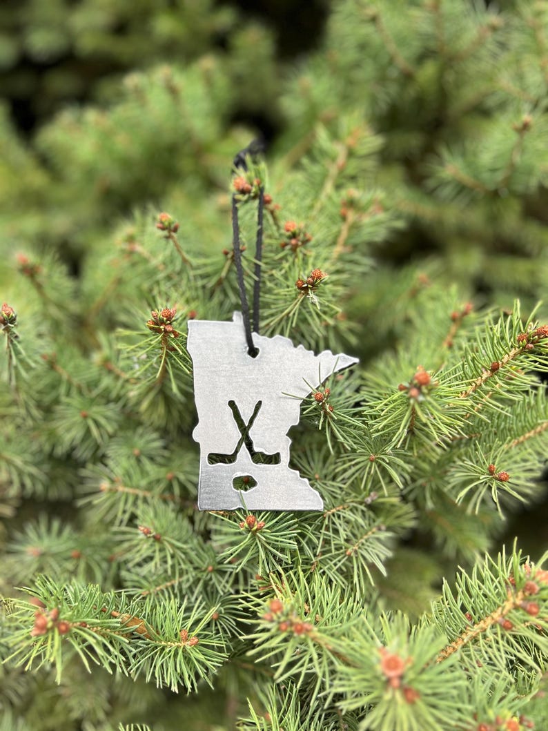 May include: A silver metal ornament shaped like the state of Minnesota with a hockey stick and puck design. The ornament is hanging on a green Christmas tree.