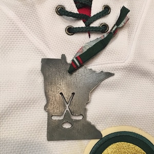 May include: A metal Minnesota state outline ornament with a hockey stick and puck design. The ornament is hanging on a white hockey jersey with green and red accents. The jersey has an NHL logo on the front.