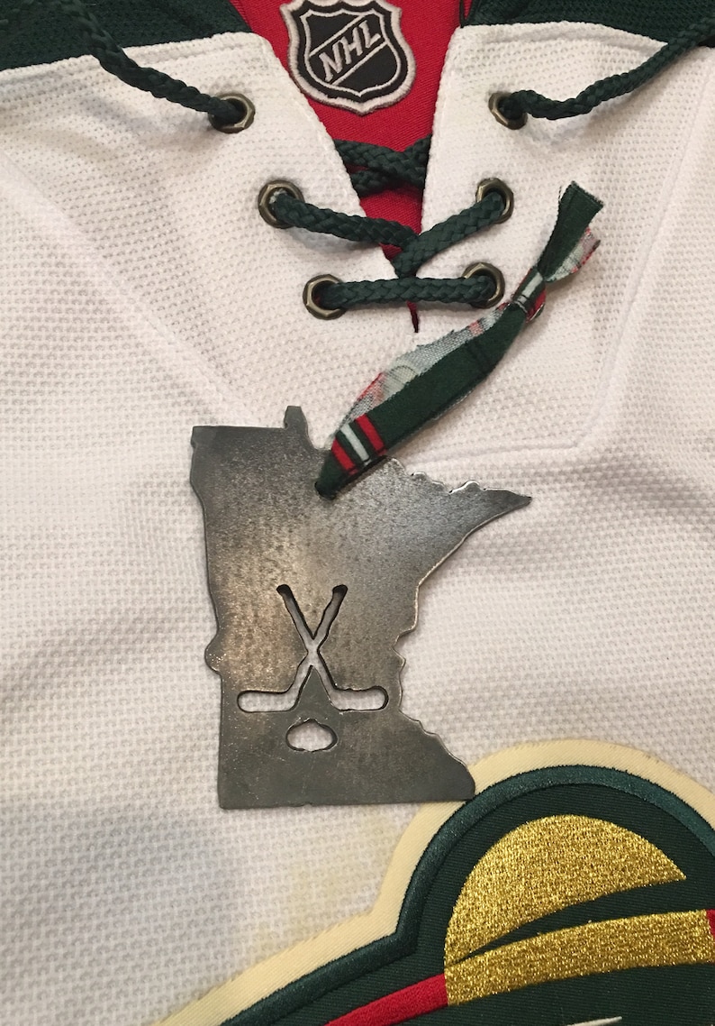 May include: A metal Minnesota state outline ornament with a hockey stick and puck design. The ornament is hanging on a white hockey jersey with green and red accents. The jersey has an NHL logo on the front.