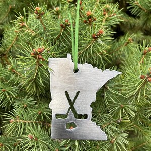 May include: A metal ornament shaped like the state of Minnesota with a hockey stick and puck design. The ornament is hanging on a green Christmas tree.