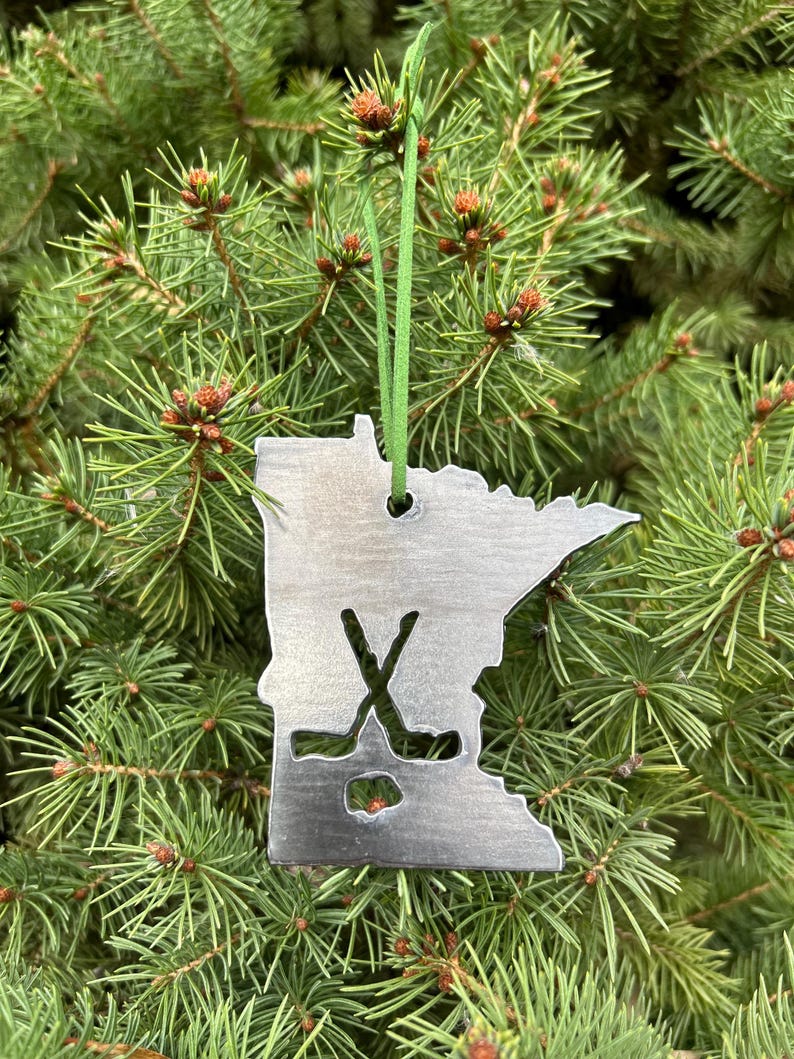 May include: A metal ornament shaped like the state of Minnesota with a hockey stick and puck design. The ornament is hanging on a green Christmas tree.