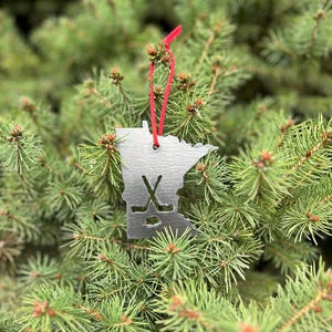 May include: A silver metal ornament shaped like the state of Minnesota with a hockey stick and puck design. The ornament is hanging from a red ribbon on a green evergreen tree.