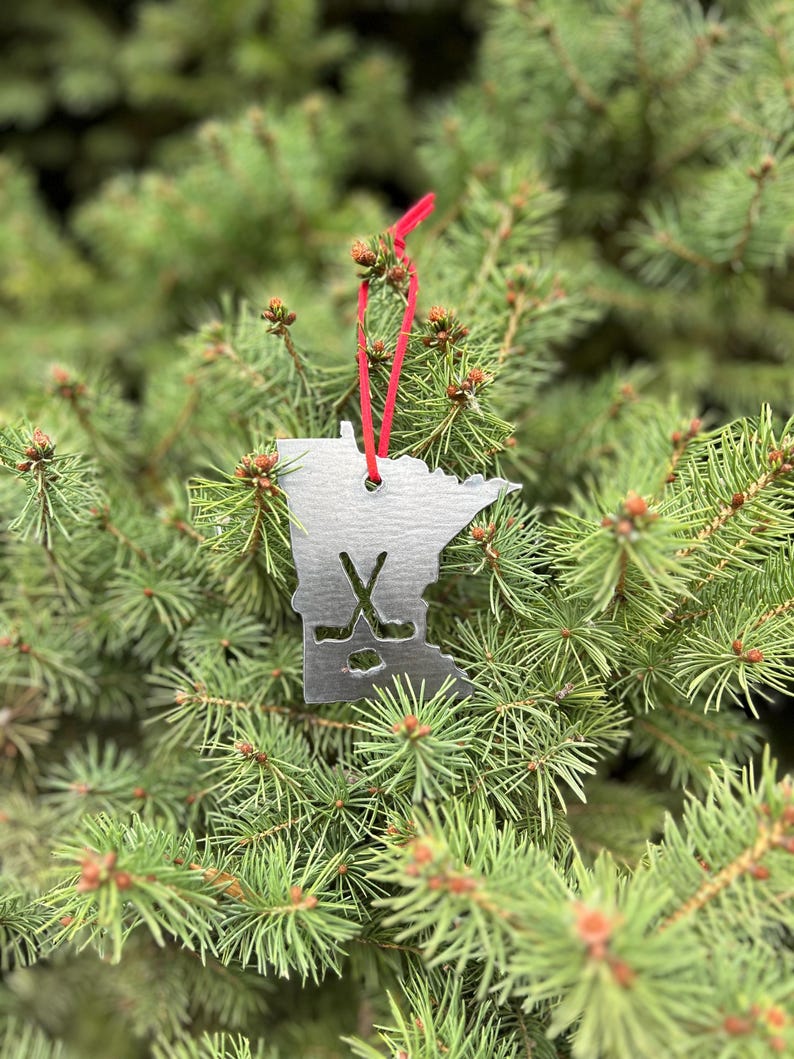 May include: A silver metal ornament shaped like the state of Minnesota with a hockey stick and puck design. The ornament is hanging from a red ribbon on a green evergreen tree.