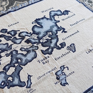 May include: A beige throw blanket featuring a detailed map of Lake Minnetonka, with blue lake outlines and city names. The blanket includes text detailing the lake's size and dimensions.
