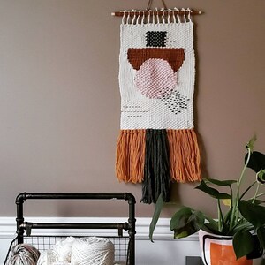 May include: A white woven wall hanging with geometric shapes in pink, brown, and black. The hanging has a fringe of orange and dark green yarn. The hanging is on a light brown wall next to a metal wire basket filled with yarn balls.