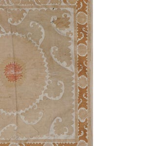 May include: Close-up of a section of a beige rug with intricate patterns. The rug features a central floral design in red and white, surrounded by white and brown decorative elements. The border is a repeating brown design.
