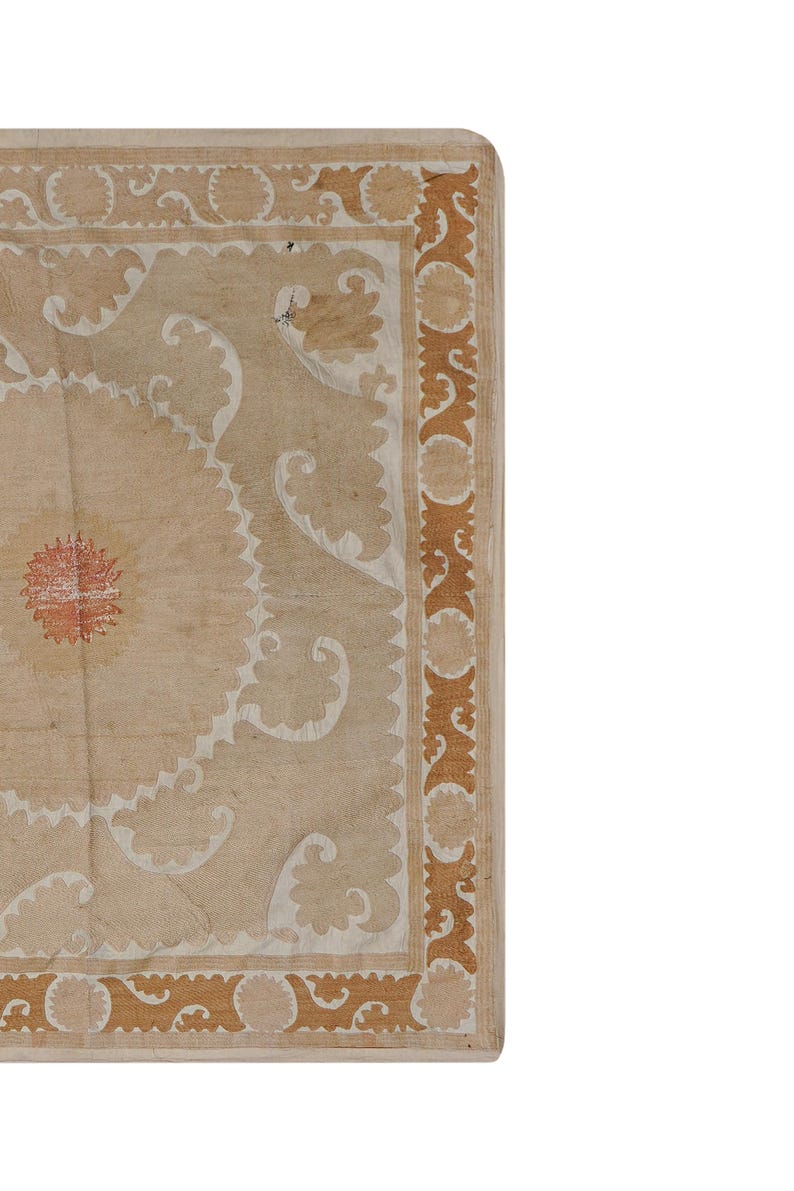 May include: Close-up of a section of a beige rug with intricate patterns. The rug features a central floral design in red and white, surrounded by white and brown decorative elements. The border is a repeating brown design.