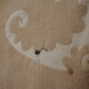 May include: Close-up of a beige textile with a textured, raised pattern. The design features large, stylized floral or cloud-like shapes in a lighter beige color, creating a contrast against the darker background. There are some dark spots on the textile.