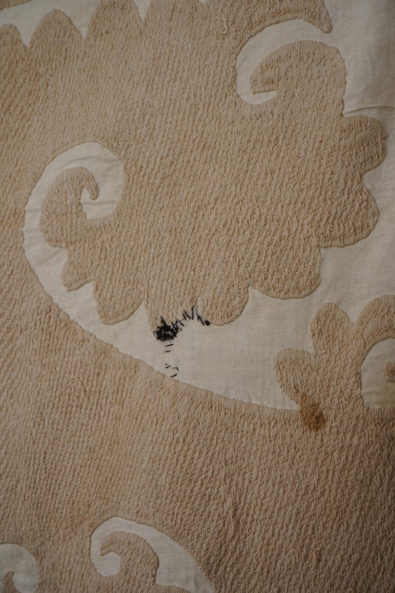 May include: Close-up of a beige textile with a textured, raised pattern. The design features large, stylized floral or cloud-like shapes in a lighter beige color, creating a contrast against the darker background. There are some dark spots on the textile.