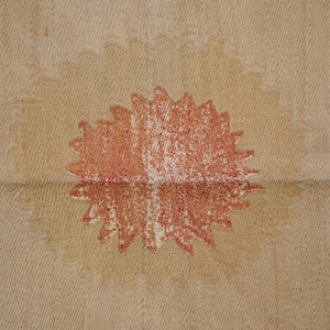 May include: A close-up of a beige fabric with a large, stylized, red and white sunburst design in the center. The sunburst has a textured, crayon-like appearance, and the fabric has a subtle, woven pattern.
