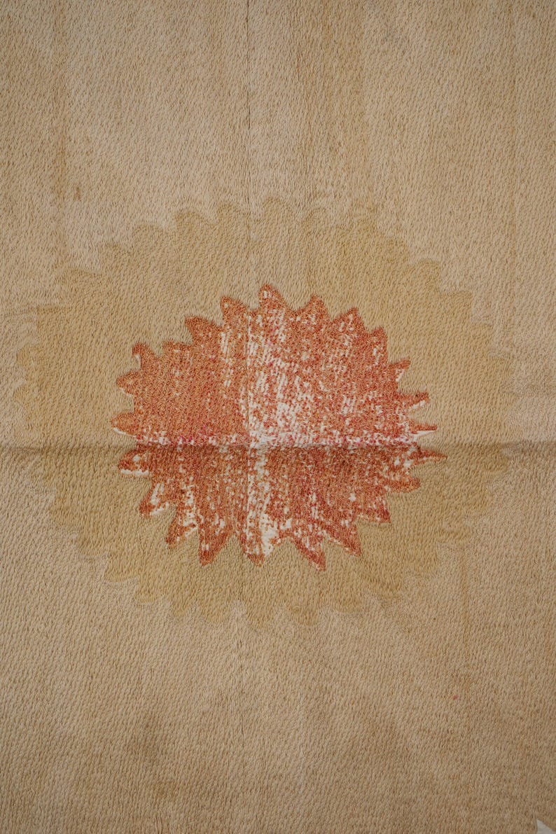 May include: A close-up of a beige fabric with a large, stylized, red and white sunburst design in the center. The sunburst has a textured, crayon-like appearance, and the fabric has a subtle, woven pattern.