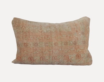 May include: A rectangular throw pillow featuring a faded, muted tan and light orange pattern. The design consists of intricate, repeating geometric and floral motifs reminiscent of a vintage rug, covering the entire surface of the pillow.