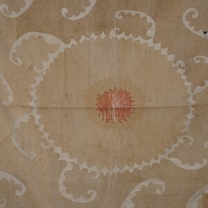 May include: A beige textile with a central circular design featuring a red center and white scalloped edges. White, swirling patterns and a brown border with circular motifs complete the design. The textile has a vintage, handcrafted appearance.