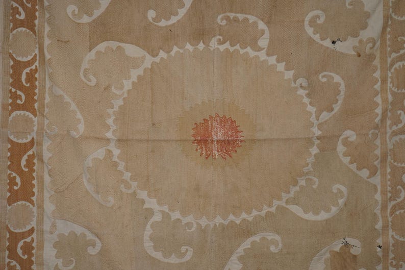 May include: A beige textile with a central circular design featuring a red center and white scalloped edges. White, swirling patterns and a brown border with circular motifs complete the design. The textile has a vintage, handcrafted appearance.