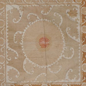 May include: A beige textile with a central circular design and ornate, repeating patterns. The center features a red and orange floral motif, surrounded by a white scalloped edge. The border has a repeating brown design.