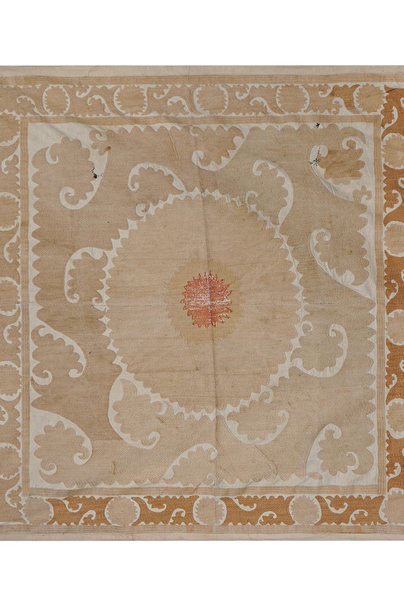 May include: A beige textile with a central circular design and ornate, repeating patterns. The center features a red and orange floral motif, surrounded by a white scalloped edge. The border has a repeating brown design.