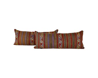 May include: Three rectangular throw pillows with a striped pattern in red, yellow, purple, and black. The pillows have a decorative tribal design. The pillows are placed on a white background.