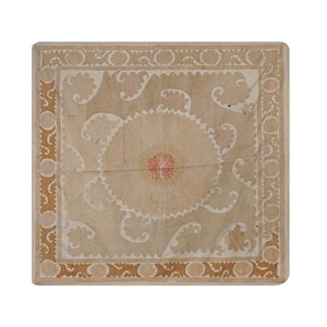 May include: Square, light-colored wooden table with a detailed, ornate design. The center features a circular pattern with red accents, surrounded by white and tan swirling motifs. The border has a repeating pattern in a darker tan color.