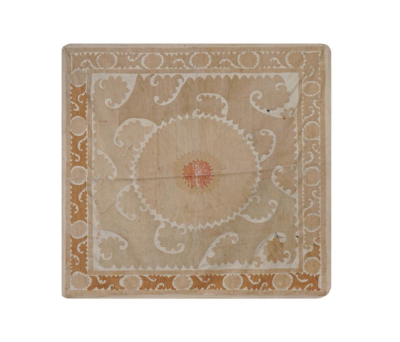 May include: Square, light-colored wooden table with a detailed, ornate design. The center features a circular pattern with red accents, surrounded by white and tan swirling motifs. The border has a repeating pattern in a darker tan color.