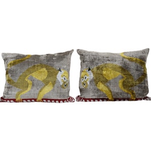 Può includere: Due cuscini decorativi in ​​velluto grigio con un motivo di scimmia ricamato giallo. I cuscini hanno un bordo rosso e bianco.