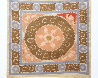 May include: A square textile wall hanging featuring a central peach-colored circular medallion with a floral motif, surrounded by a brown ring of stylized leaf patterns, a light blue border, and an outer frame of brown and blue floral designs.