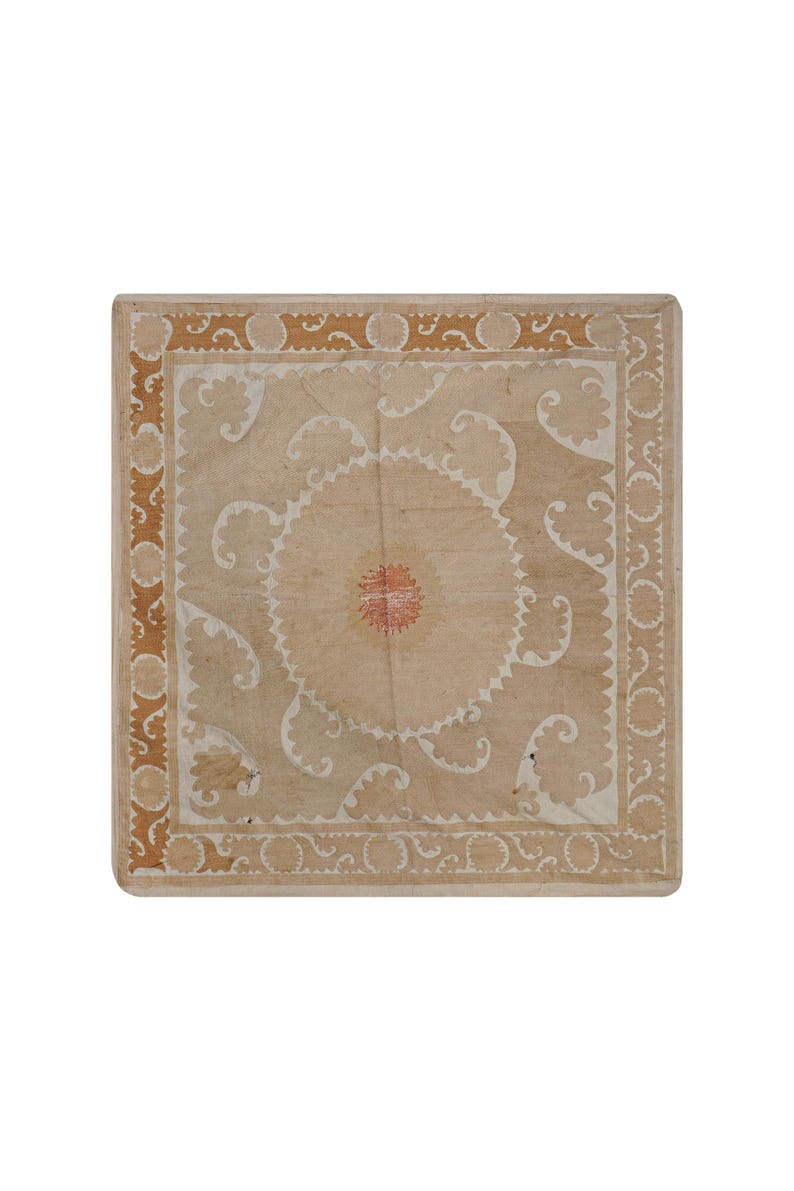 May include: A square, beige textile with a central circular design and ornate white and brown border. The center features a red and white floral pattern, surrounded by white, curved embellishments.