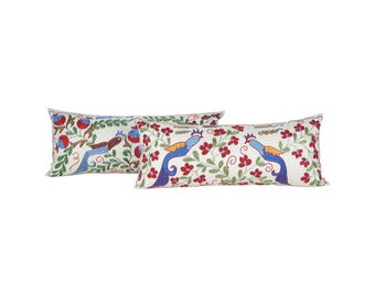 May include: Two rectangular decorative pillows with embroidered designs. One pillow features a bird with a long tail, red berries, and green leaves. The other pillow has two blue birds with red flowers and green leaves on a white background.